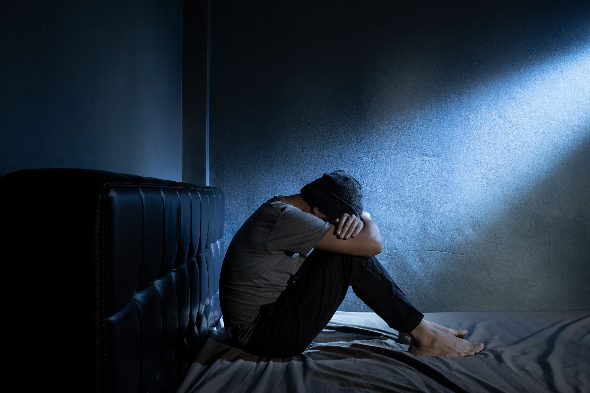 A person sits on a bed in a dimly lit room, hugged knees, and head down. Soft blue light from a single source creates a somber, reflective mood.