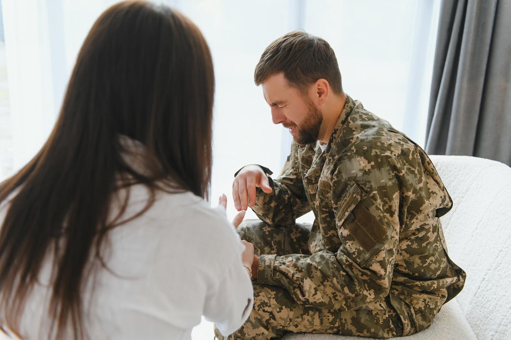 A person in military camouflage sits on a couch, engaged in conversation with another person facing him. The setting is calm and supportive.