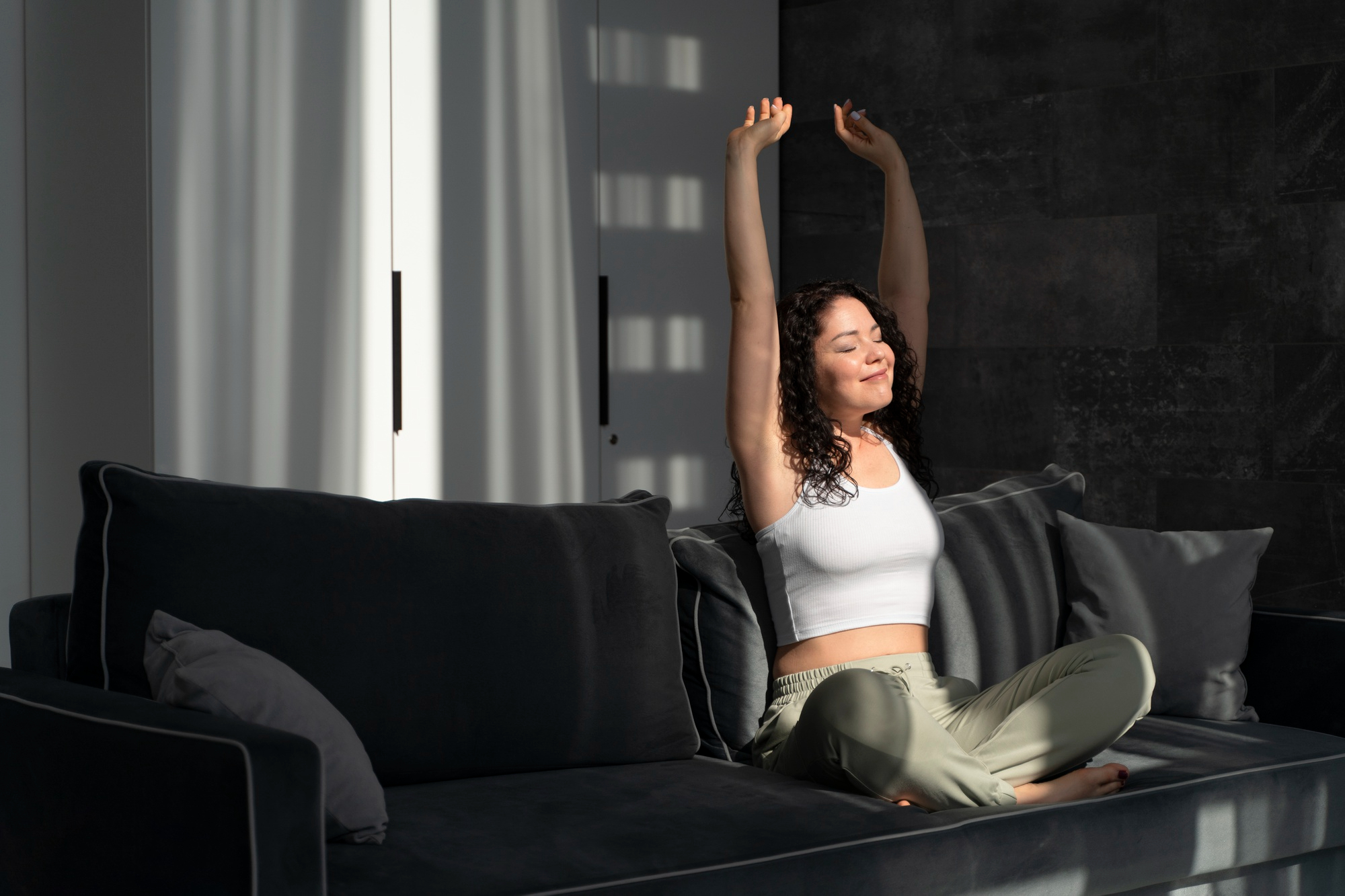 Woman in white top and green pants sits cross-legged on a dark couch, stretching with arms raised, sunlight casts soft shadows, creating a serene mood.