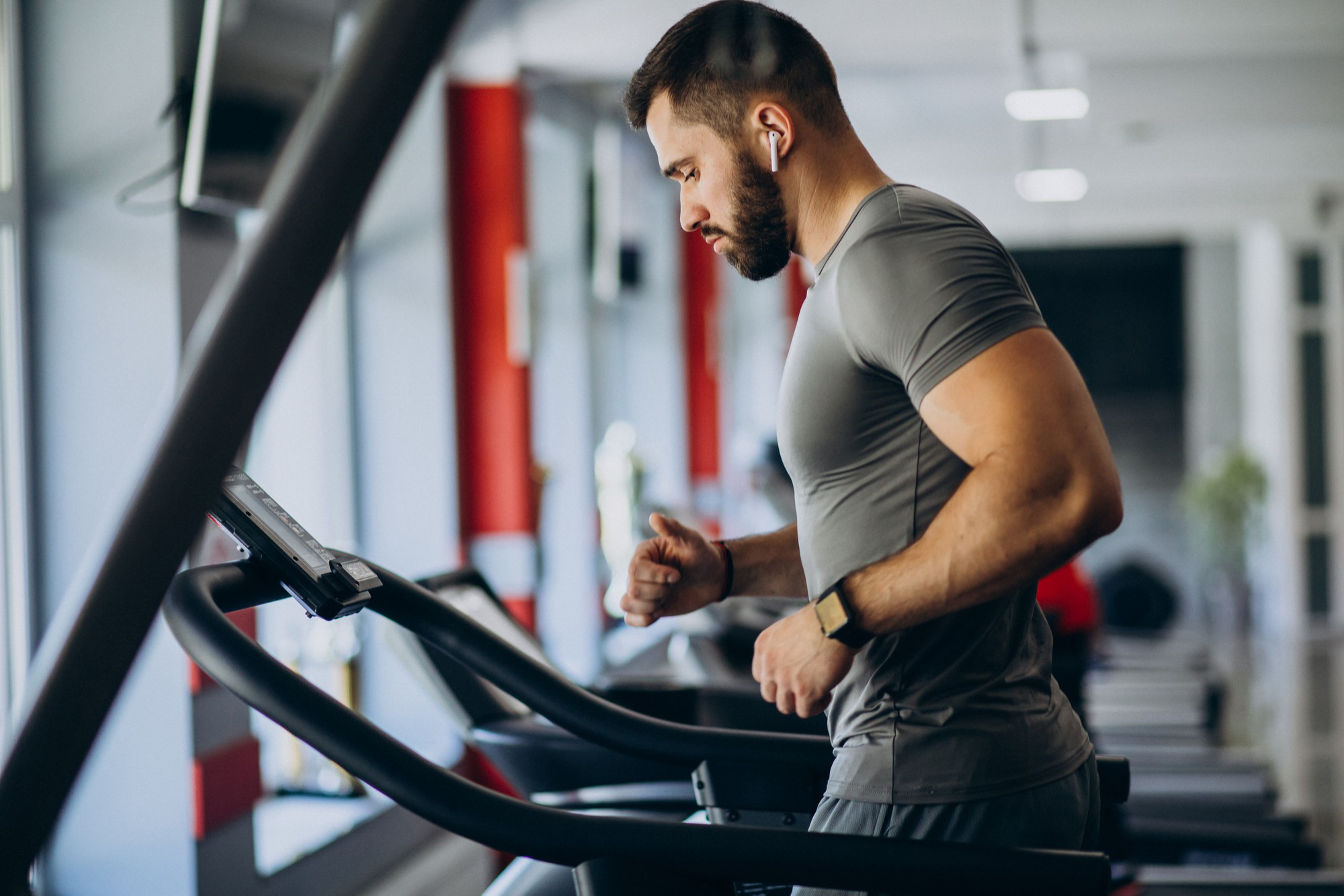Man running on a treadmill in a gym, wearing a gray shirt and earbuds. The space is modern and brightly lit, conveying focus and determination.