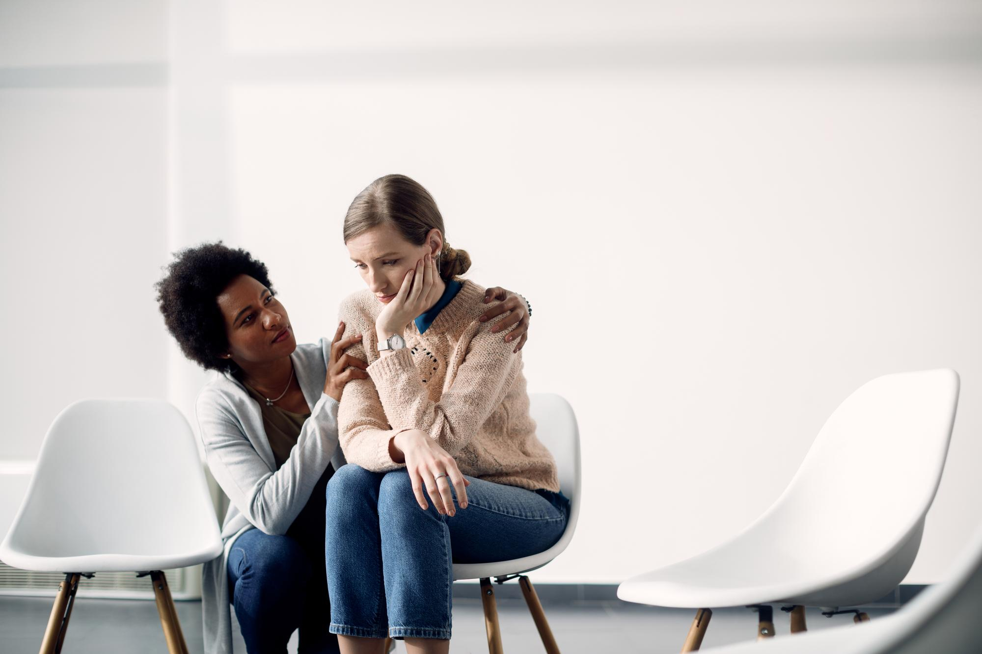A woman consoles a distressed friend seated on a white chair in a bright room. She offers comfort with a gentle hand on her shoulder, creating a supportive tone.