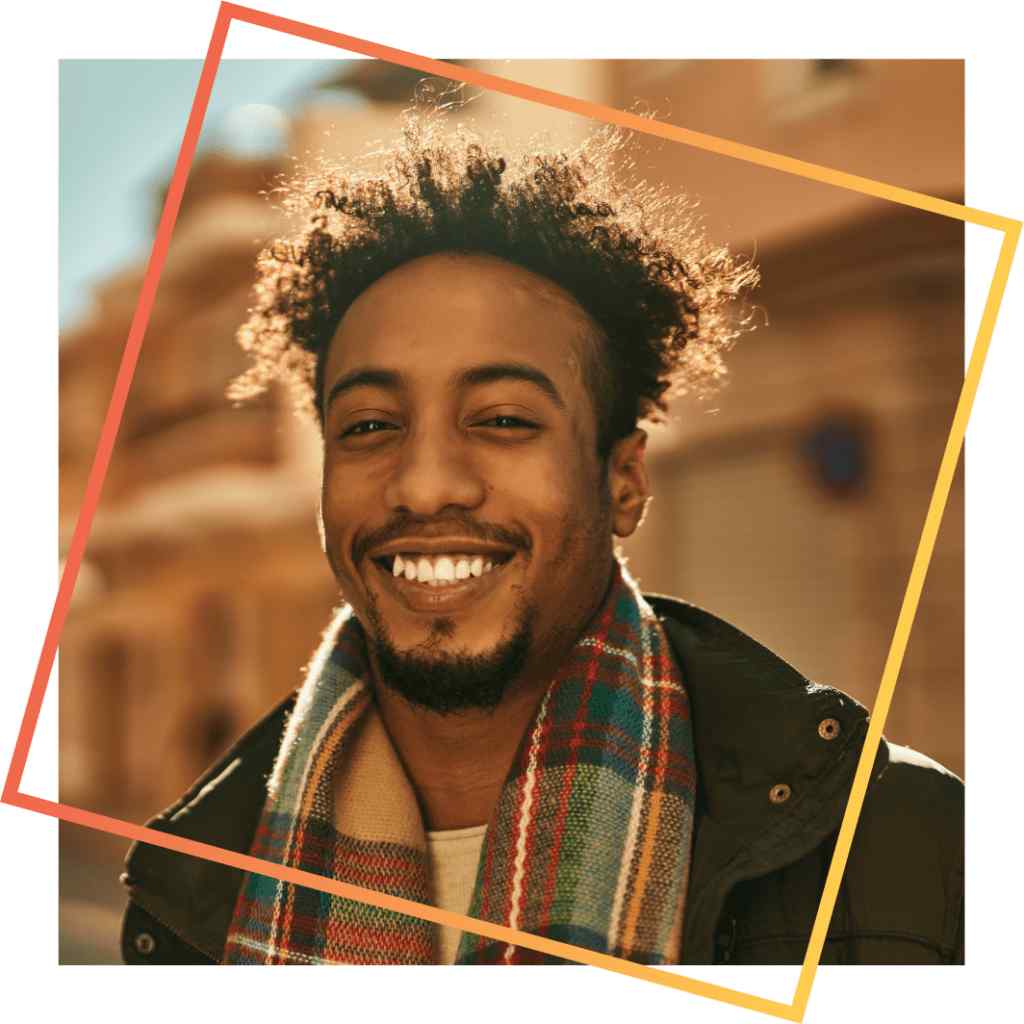 Smiling man with curly hair, wearing a colorful plaid scarf and dark jacket, standing outdoors in soft sunlight with blurred buildings in the background.